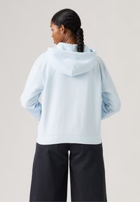 Ανοιχτό γαλάζιο φούτερ με κουκούλα, με άνετη εφαρμογή και ριμπ τελείωμα. Φορεμένο με σκούρο, φαρδύ παντελόνι, παρουσιάζοντας έναν casual στιλ.