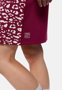 Dunkelroter Sportrock mit weißem Leopardenmuster auf einer Seite, aus elastischem Stoff; Logoschild in der Nähe des Saums sichtbar.