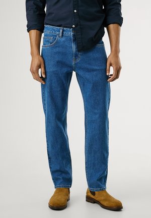 Mand iført blå jeans, sort skjorte og lysebrune ruskindsstøvler, stående mod en ensfarvet lys baggrund.