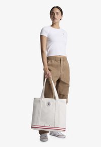 Platnena torba u bijeloj boji s crvenim prugama, čvrstim drškama i okruglom oznakom. Model nosi usku bijelu majicu i kaki hlače.