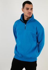 Sudadera de forro polar azul con cuello alto, cierre de cremallera y puños acanalados. Presenta un detalle en el panel frontal, textura suave y corte holgado.