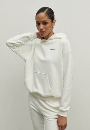 Kobieta z włosami zaczesanymi do tyłu, ubrana w biały kaptur i pasujące dresy, oba z napisem „LAMANIA”, pozująca z jedną ręką za szyją.