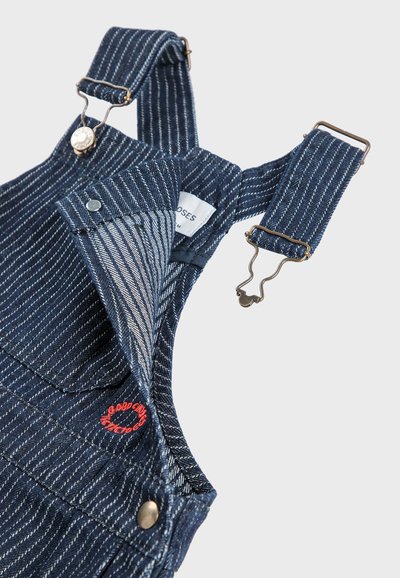 Salopette en denim rayée bleue avec des clips en métal réglables, poche avant et petit détail de logo rouge brodé au niveau de la poitrine.