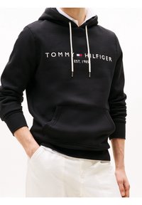 Black hoodie featuring a front pocket, white drawstrings, and "TOMMY HILFIGER EST. 1985" printed across the chest in contrasting lettering.