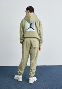 Oliven grøn hættetrøje og matchende joggingbukser lavet af blødt materiale, med en grafisk print på ryggen og hvide sneakers med strukturerede såler.