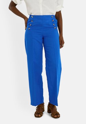 Femme portant un pantalon taille haute bleu vif à six boutons et des sandales à talons imprimé léopard marron, debout sur un fond blanc.
