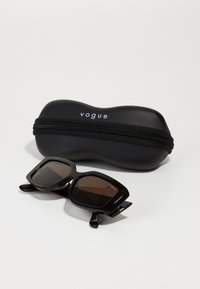 Juodos tortoiseshell saulės akinius su kvadratiniais rėmeliais, rudos atspalvio lęšiais ir "Vogue" logotipu ant šventininkų, papildytus juoda užtrauktuko dėžute.
