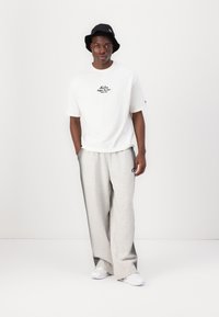 White short-sleeve t-shirt with black logo, paired with gray sweatpants and a black bucket hat. Model wears white sneakers.