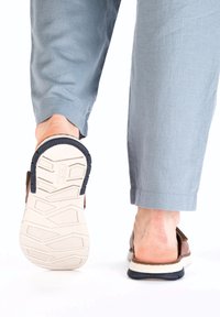 Braune Slipper mit strukturiertem Obermaterial, weißer und marineblauer Gummisohle und einem ausgearbeiteten profil. Hellblaue, locker sitzende Hose.