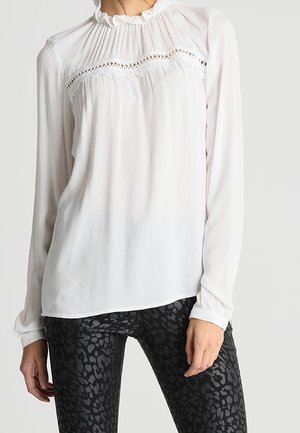 Kobieta ubrana w białą bluzkę z długim rękawem z koronkowymi zdobieniami i falbanianym kołnierzykiem, zestawioną z czarnymi, wzorzystymi, dopasowanymi spodniami.