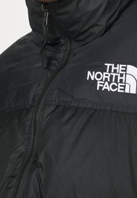 Giacca imbottita nera con collo alto, dotata di zip integrale e logo The North Face bianco sul lato sinistro del petto. Tessuto dalla texture liscia.