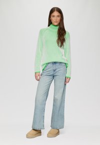 Grün-weiß gestreifter Rollkragenpullover mit langen Ärmeln, kombiniert mit hellblauen weiten Jeans und beigen Plateauschuhen.