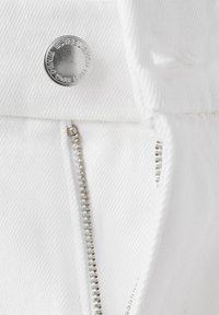 Záber zblízka na bielu denimu tkaninu so čiastočne rozopnutým strieborným kovovým zipsom a strieborným gombíkom s nápisom "MNG DENIM ESTABLISHED 1984".