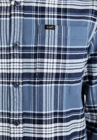 Camisa de cuadros azul hecha de tela de algodón. Presenta patrones en blanco, azul marino y azul claro, con un bolsillo en el pecho y una etiqueta de la marca. Se ven seis botones.