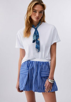 Blonde Frau, die ein weißes Kurzarmshirt, einen blau gemusterten Halsschal, einen blau gestreiften Rock mit großen Taschen und silberne Armbänder trägt, steht vor weißem Hintergrund.