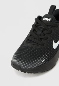 Μαύρο αθλητικό παπούτσι με λευκό λογότυπο swoosh, διάτρητο πάνω μέρος, μαύρα κορδόνια και θηλιά στο τακούνι, σε γκρίζο φόντο.