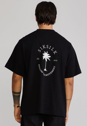 Mann trägt ein schwarzes T-Shirt mit weißem Palmenmotiv und dem Schriftzug "SIKSILK" auf dem Rücken, steht vor einem einfarbigen hellgrauen Hintergrund.