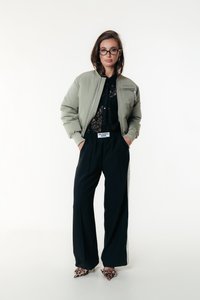 Giacca bomber verde, top in pizzo nero, pantaloni su misura neri con righe laterali e tacchi animalier. Presenta una silhouette oversize e dettagli di cucitura.