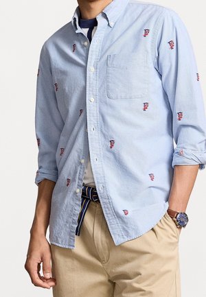 Mężczyzna ubrany w jasnoniebieską koszulę na guziki z małymi, czerwono-niebieskimi haftowanymi wzorami, beżowe spodnie, paskiem w paski i zegarkiem na nadgarstku.