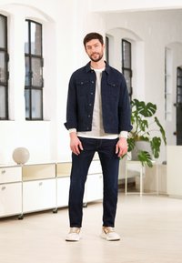 Donkerblauwe denimjack met borstzakken, lichtgrijze trui en donkerblauwe jeans. Bruin en witte sneakers. Binnenomgeving met ramen en planten.