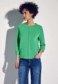 Maglia verde a maniche lunghe con scollatura tonda e dettaglio a pieghe, abbinata a jeans in denim blu. Il tessuto appare liscio con una leggera lucentezza.