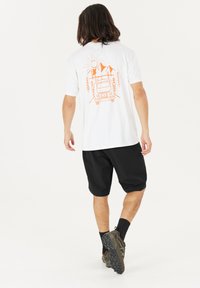 Witte T-shirt met oranje afbeelding van een bus, zon en bergen op de achterkant, gecombineerd met zwarte shorts en gestructureerd, donker schoeisel.