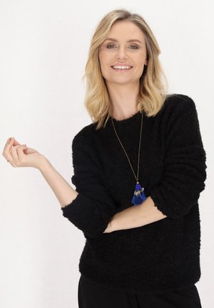 Un pull noir en tricot duveteux avec des poignets larges. Accessoirisé avec une chaîne en or ornée d'un pendentif à pompon bleu.