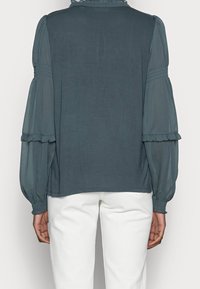 Femme portant un blouse à manches longues bleu sarcelle foncé avec des manches transparentes et bouffantes, vue de dos sur un fond uni.