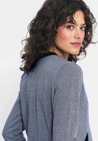 Kobieta z kręconymi ciemnymi włosami, ubrana w niebiesko-białą kurtkę w deseń jodełki, spogląda przez ramię na tle jednolitego, białego tła.