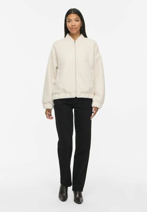 Femme portant une veste polaire blanche zippée, un pantalon noir et des bottines noires, debout sur un fond blanc uni.