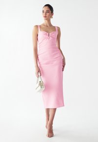 Robe midi rose avec une silhouette ajustée, décolleté carré et fronces au centre. Accompagnée d'un petit sac à main blanc.