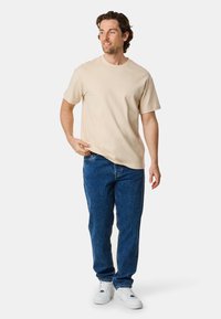 Beige short-sleeve t-shirt with a round neckline, paired with blue denim jeans and white sneakers. Casual fit, smooth texture, and clean design.