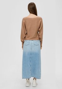 Brauner gerippter Pullover mit langen Ärmeln, kombiniert mit einem hellblauen Denim-Maxikleid mit aufgesetzten Taschen und leicht fransigem Saum. Weiße Sneakers.
