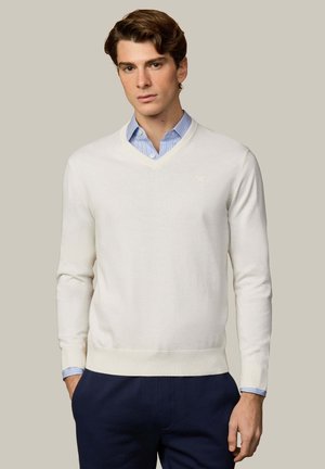 Jonge man die een wit V-hals trui draagt over een lichtblauw gestreept overhemd, gecombineerd met donkerblauwe pantalon, staand tegen een beige achtergrond.