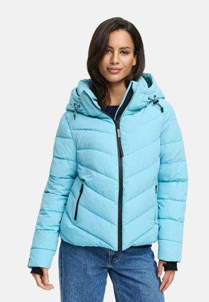 Donna che indossa una giacca a vento imbottita di colore blu chiaro con cappuccio e cerniera nera, abbinata a jeans blu, su uno sfondo bianco.