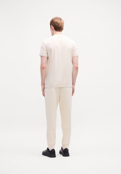 T-shirt en coton beige associé à un pantalon de survêtement crème. Texture lisse, manches courtes et poignets élastiques aux chevilles. Fini avec des baskets noires.