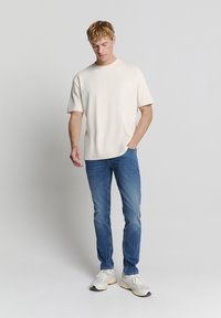 Korte mouw crèmekleurig T-shirt met eenRelaxed fit, gecombineerd met blauwe slim-fit jeans en grijze sneakers, staand tegen een lichte achtergrond.