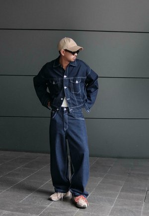 Man wearing dark denim jacket and wide-leg jeans, beige cap, sunglasses, and sneakers, standing against a plain gray wall.