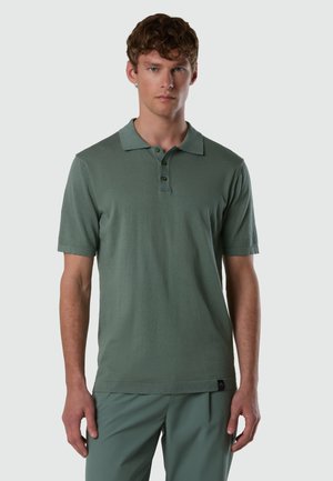 Giovane uomo con capelli corti che indossa una polo a maniche corte verde smorzato e pantaloni verdi abbinati, in piedi davanti a uno sfondo grigio uniforme.