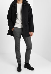 Černá nepromokavá parka s kapucí, s kapsami na zip a texturovaným bílým svetrem pod ní. Šedé kostkované kalhoty a černé boty.