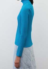 Turquoise ribgebreide coltrui met lange mouwen, nauwsluitend ontwerp en gestructureerd patroon. Draagt een lichtgrijze doorgestikte rok.