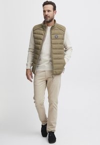 Olivenfarbener Steppweste mit Reißverschluss und Logo-Patch. Trägt man über einem leichten Pullover. Helle beige Hose und schwarze Schuhe runden das Outfit ab.