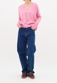 Różowy sweter z dekoltem w serek, z prążkowanymi mankietami i hemem, zestawiony z luźnymi, ciemnoniebieskimi dżinsami oraz brązowymi butami sznurowanymi.