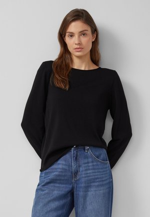 Frau mit langen braunen Haaren, die ein schwarzes Langarmshirt und blaue Jeans trägt, vor einem schlichten hellgrauen Hintergrund stehend.