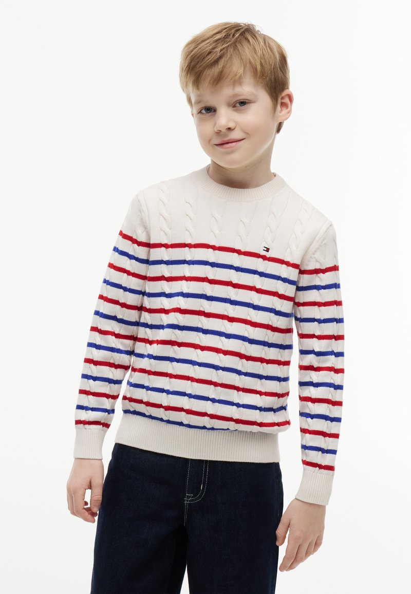 Garçon blond portant un pull en maille torsadée blanc avec des rayures horizontales rouges et bleues et un pantalon bleu foncé, debout devant un fond blanc.