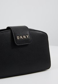 Clutch in pelle nera con trama, dotato di chiusura a patta e lettere dorate che formano "DKNY", su uno sfondo chiaro e semplice.