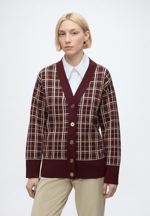 Kvinde med kort blondt hår iført en vinrød ternet knap-op cardigan over en hvid skjorte med krave og beige bukser mod en ensfarvet hvid baggrund.