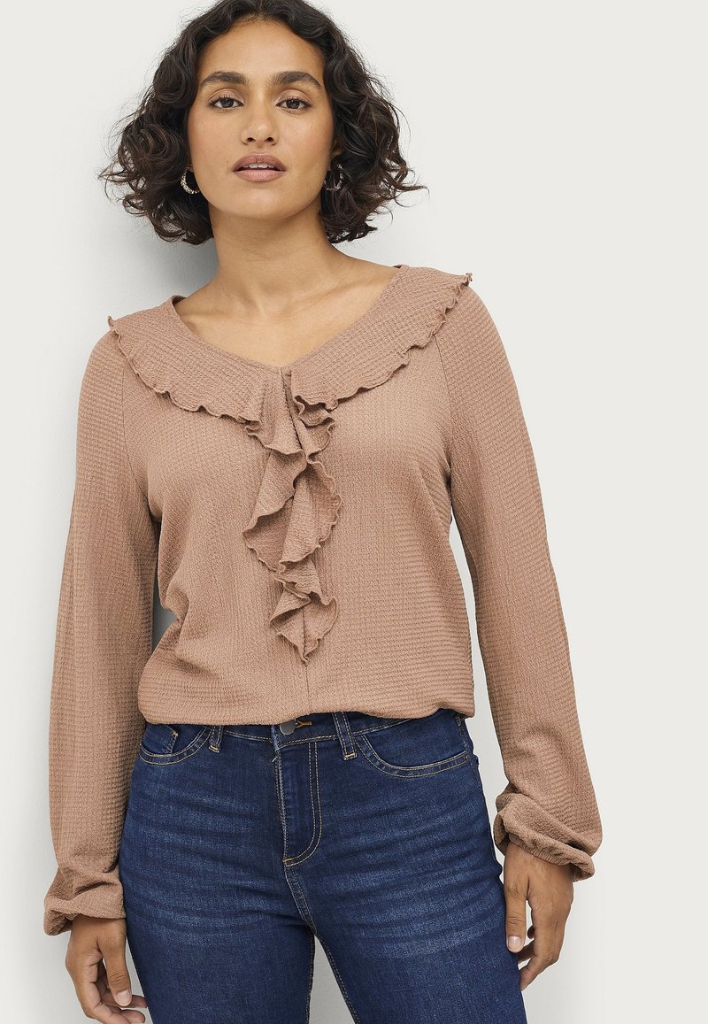 Langarm, V-hals top van zacht, gestructureerd lichtbruin stof, met een ruches bij de halslijn en knoopsluiting, gecombineerd met donkerblauwe jeans.