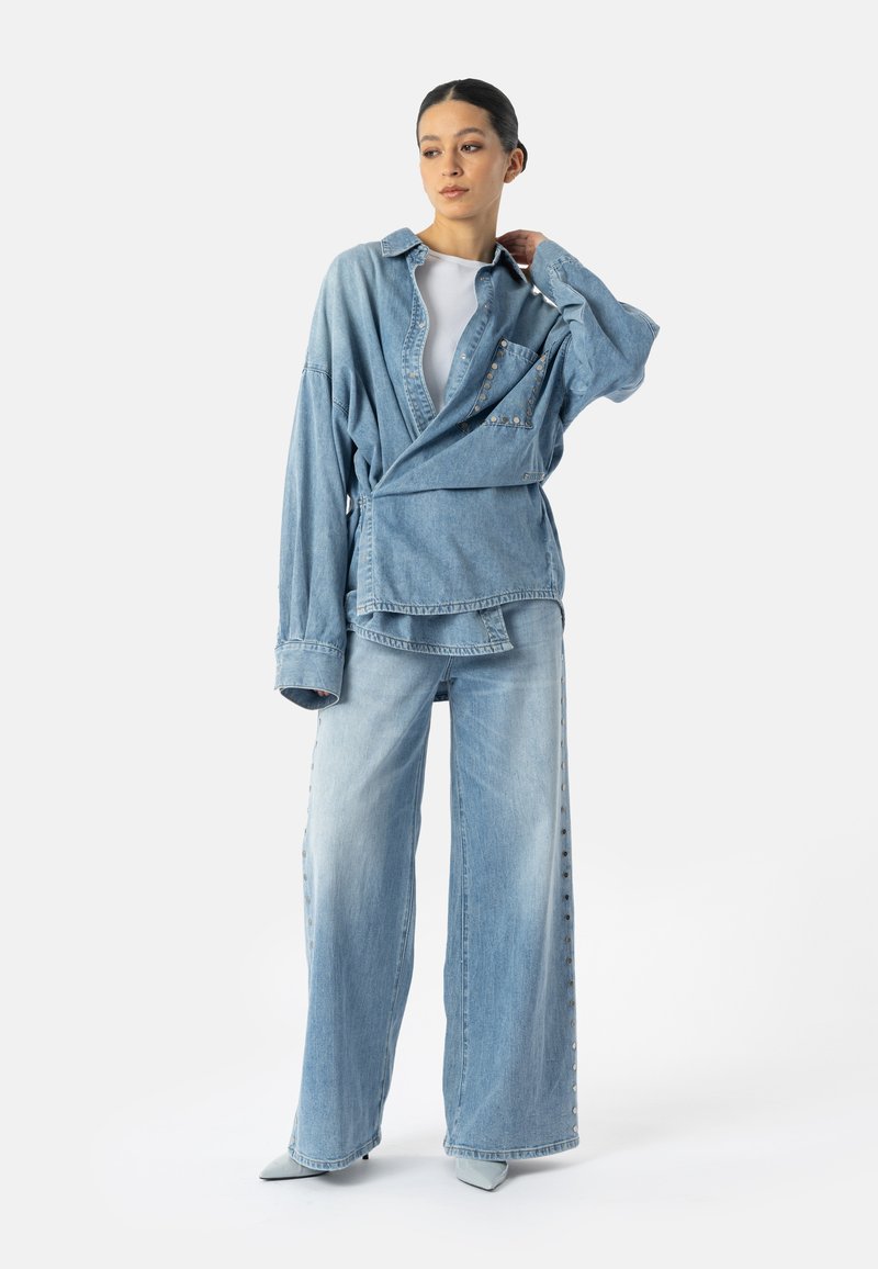 Femme portant une veste en jean bleu clair oversize et un jean à jambes larges avec des clous sur les côtés, posant avec la main touchant le cou, sur fond blanc.