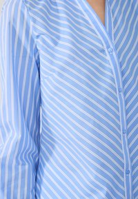 Light blue button-up shirt with white vertical stripes, long sleeves, and a pocket on the left side. Smooth texture and classic collar design.